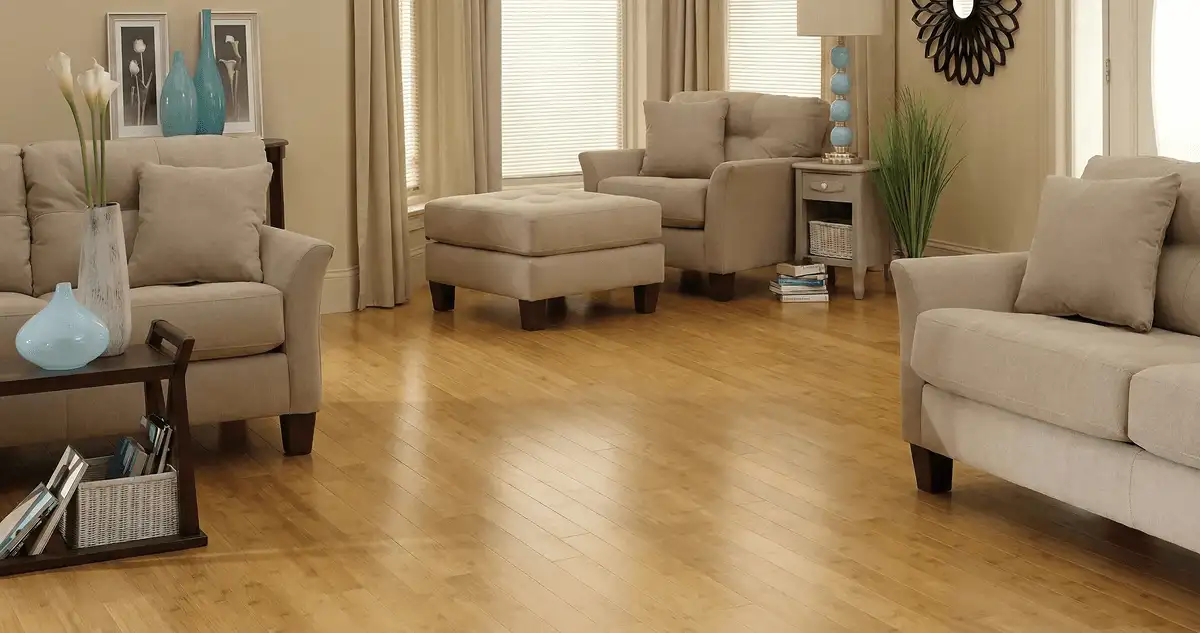A living room with professionally installed bamboo flooring, beige sofas, a matching ottoman, a side table with a blue lamp, vases, framed pictures, and large windows with blinds and curtains in Newark, NJ